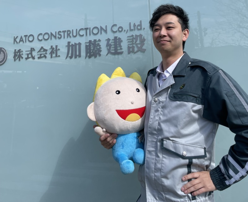 株式会社加藤建設の臼井さんがゆるキャラのぬいぐるみを持って社名前で撮影する様子