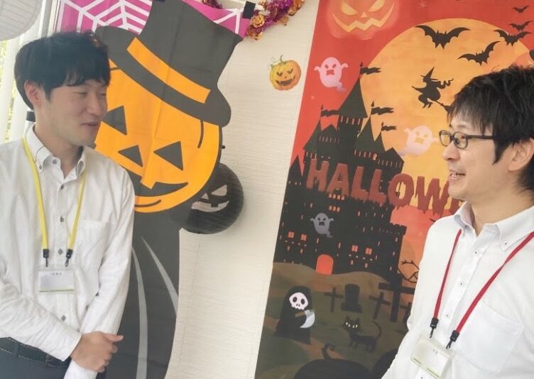 ハロウィンのPOP類の前で話す株式会社アルファの若手社員